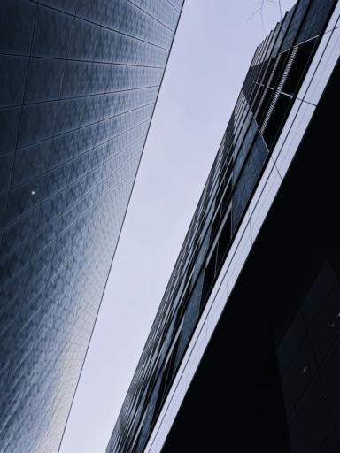 Low-angle view of two modern buildings with reflective glass surfaces set against a cloudy sky. The buildings create a sharp, angular perspective converging towards the center of the image. Subtle patterns can be seen on the facade of the left building, with both structures appearing dark and sleek.