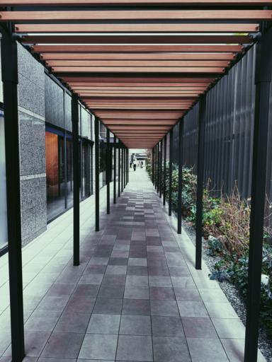 A long, covered walkway with a wooden slatted roof and metal supports stretches between two buildings. The floor is paved with square tiles, and there are small plants lining the right side with gravel. The scene leads to an open area where two people are seen in the distance.